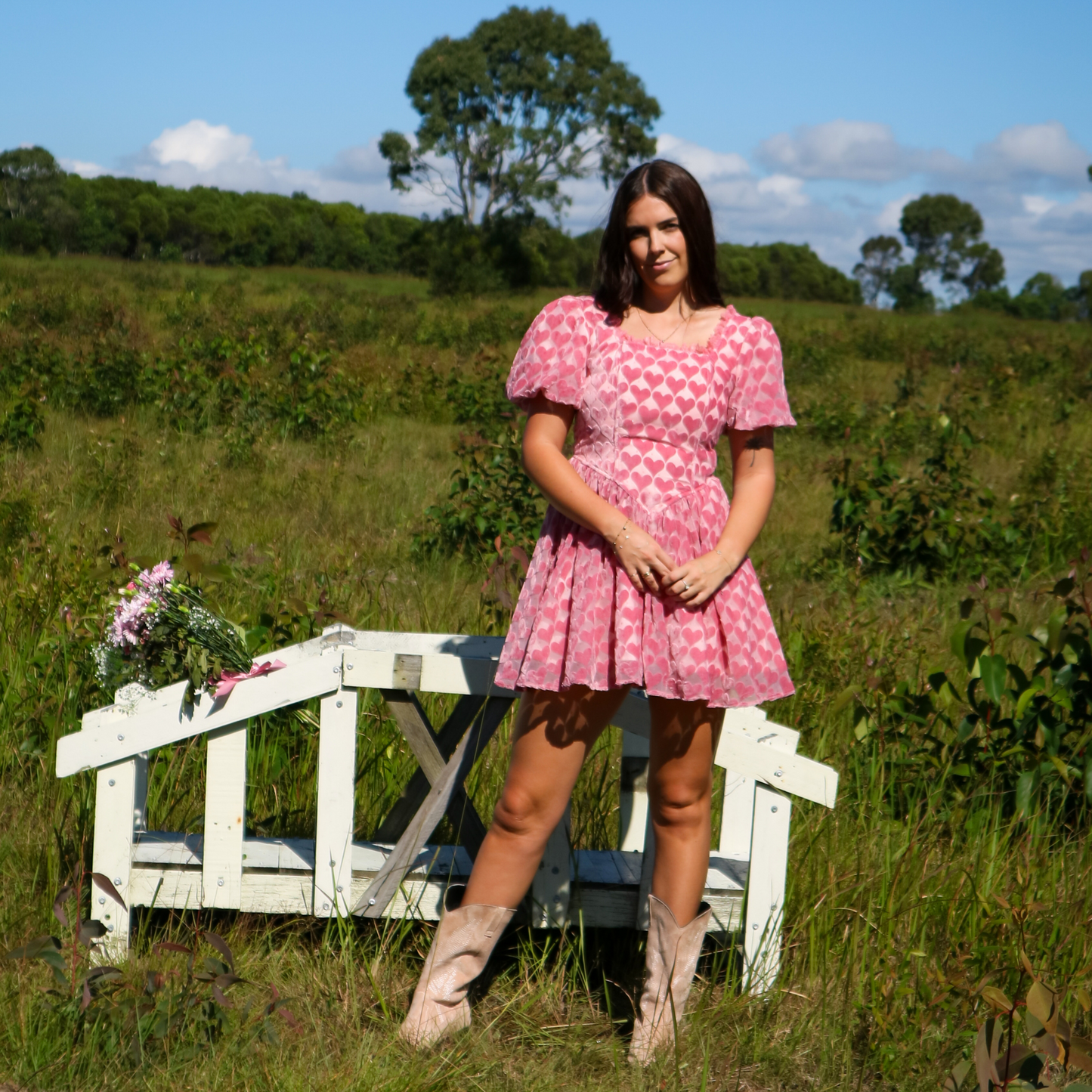 Woman in a pink heart dress standing next to a white bench in a grassy field.