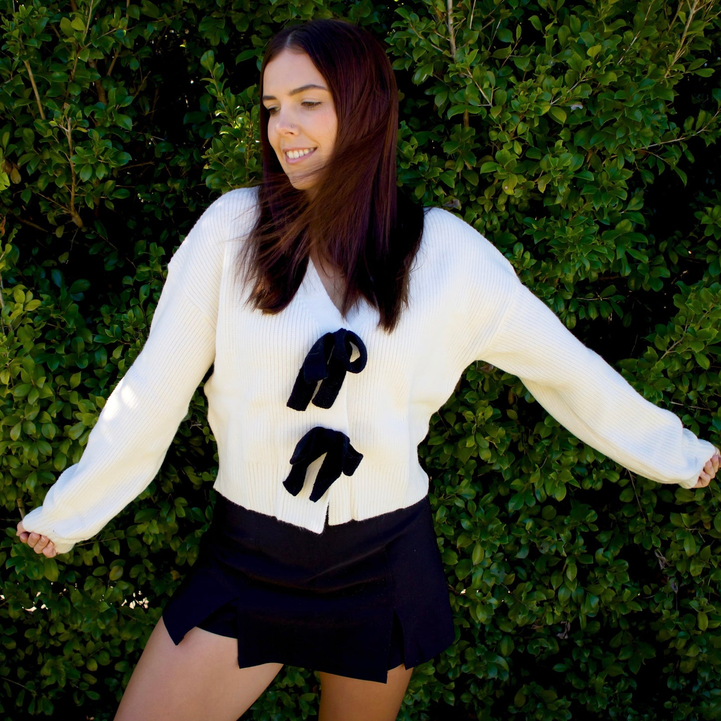 Woman wearing a white sweater with black bow ties in front of green foliage