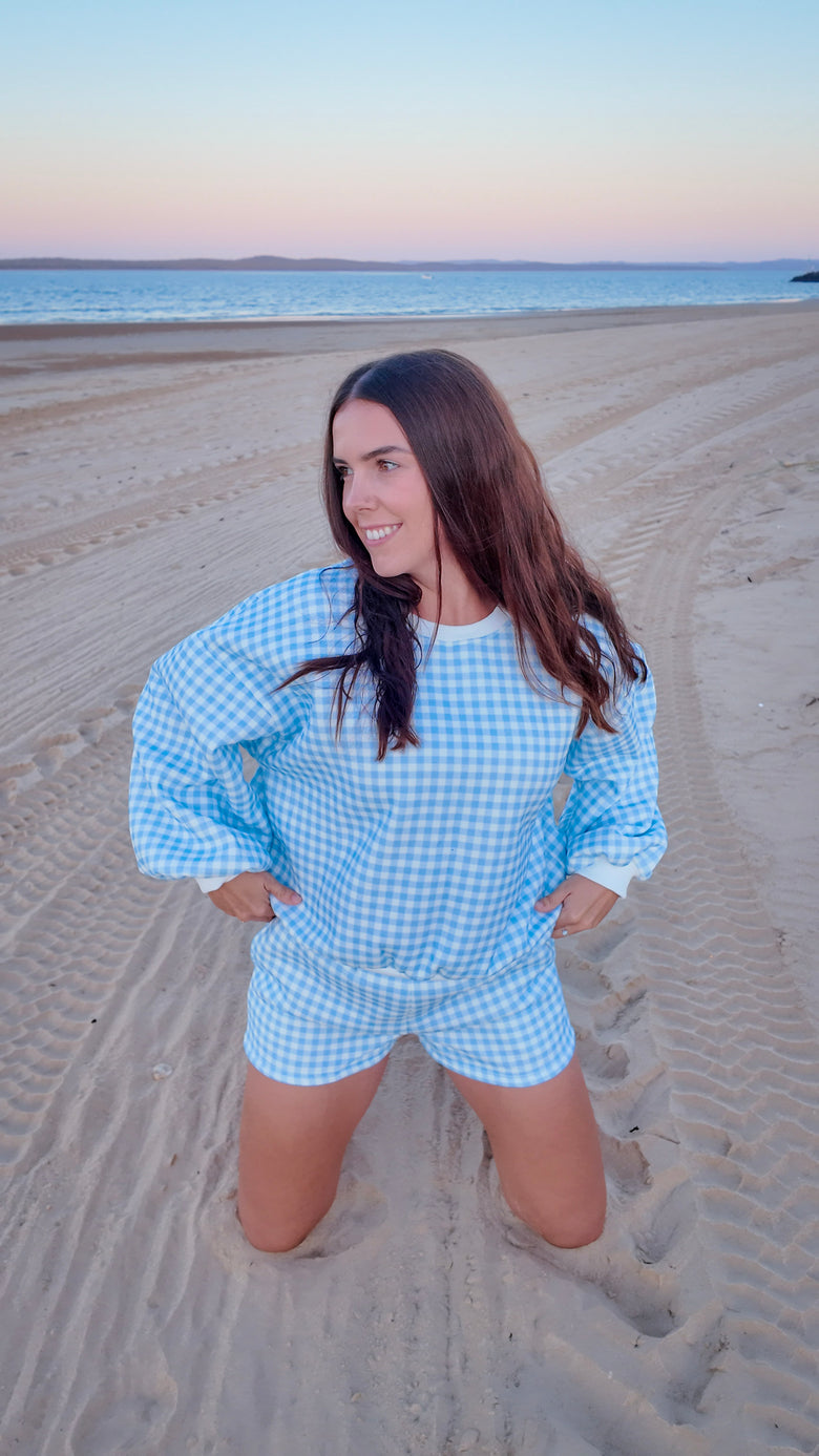 Woman in a blue checkered outfit standing on a sandy beach with ocean in the background