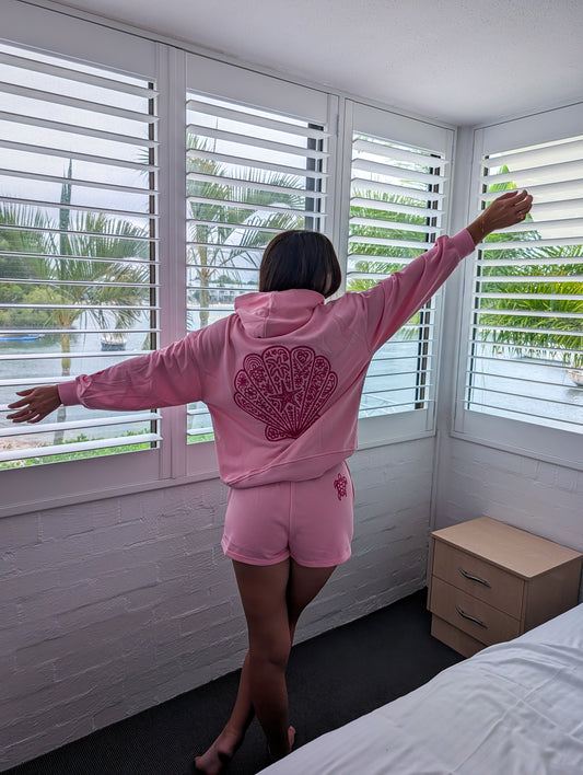 Person wearing a pink hoodie with a design on the back, standing in a room with white shutters.