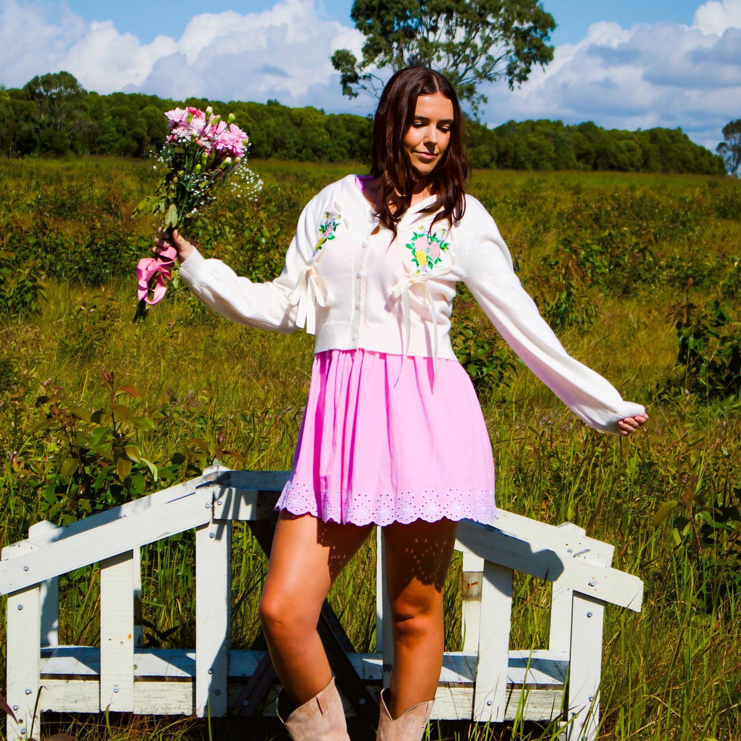 Woman in a white cardigan standing on a wooden bridge in a field with flowers.