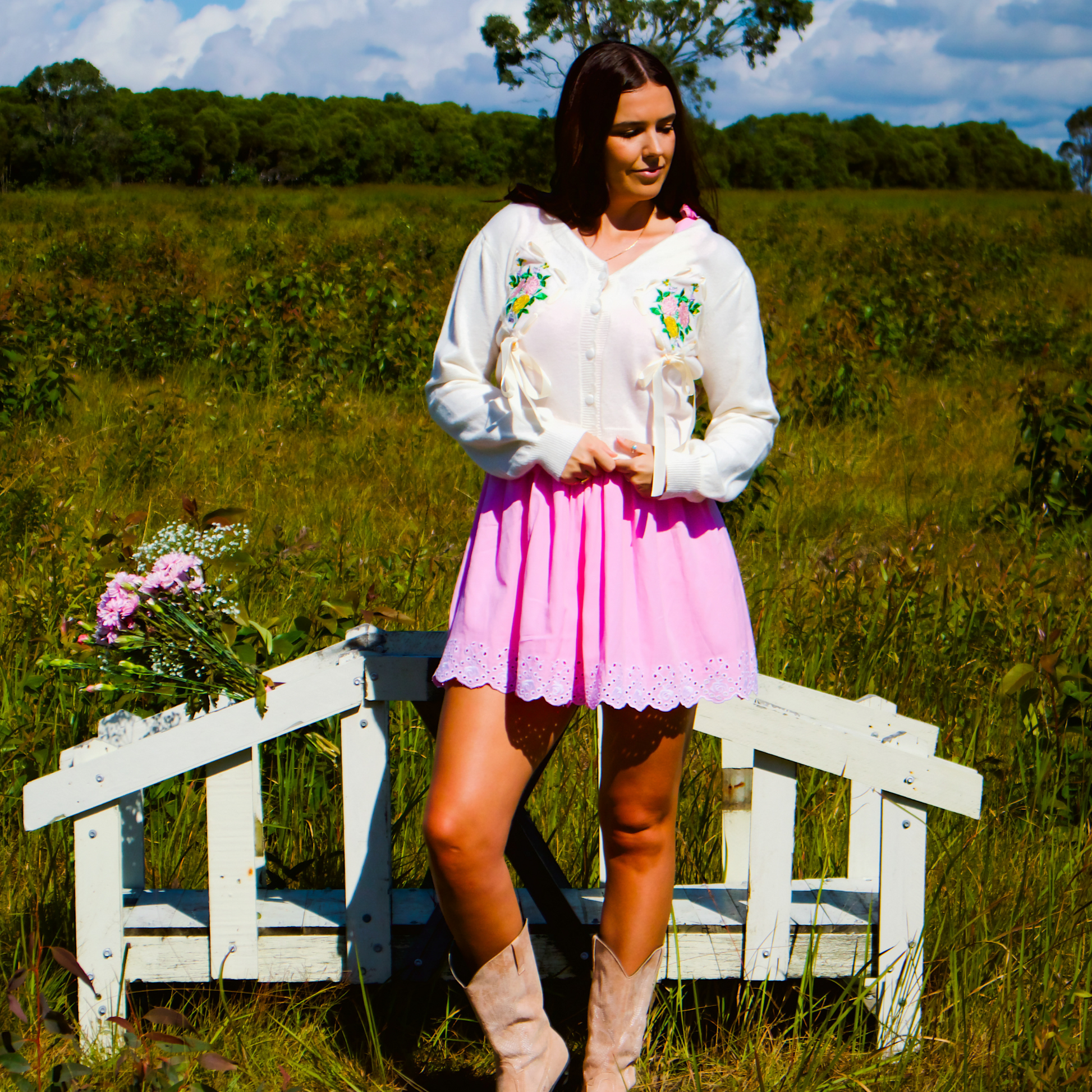 Woman in a white cardigan with floral embroidery standing in a field with a white bench.