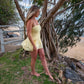 Woman in a yellow dress standing near a large tree on a beach.