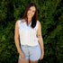 Woman wearing a white sleeveless top and denim shorts standing in front of green foliage.