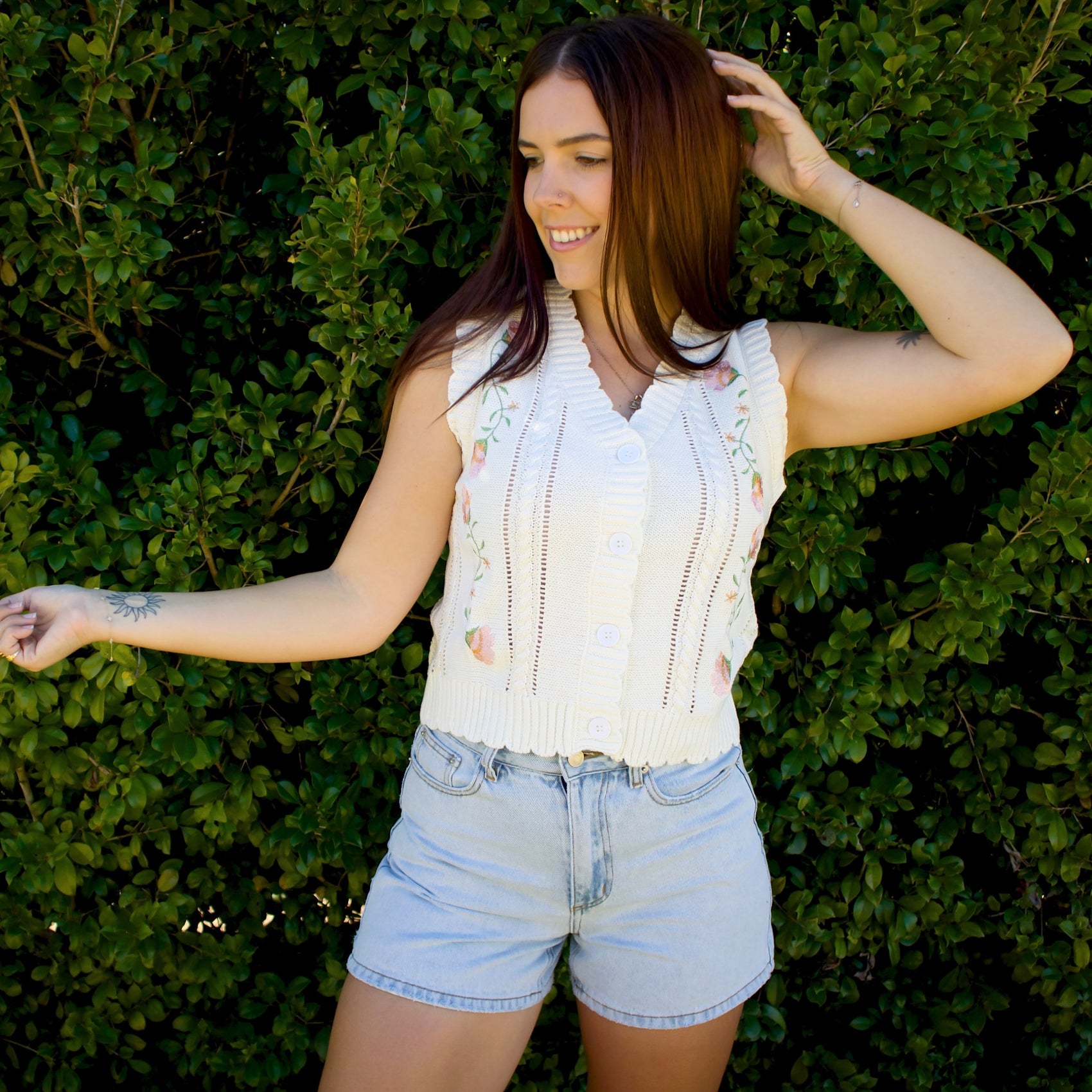 Woman wearing a white sleeveless top and denim shorts standing against a green hedge.
