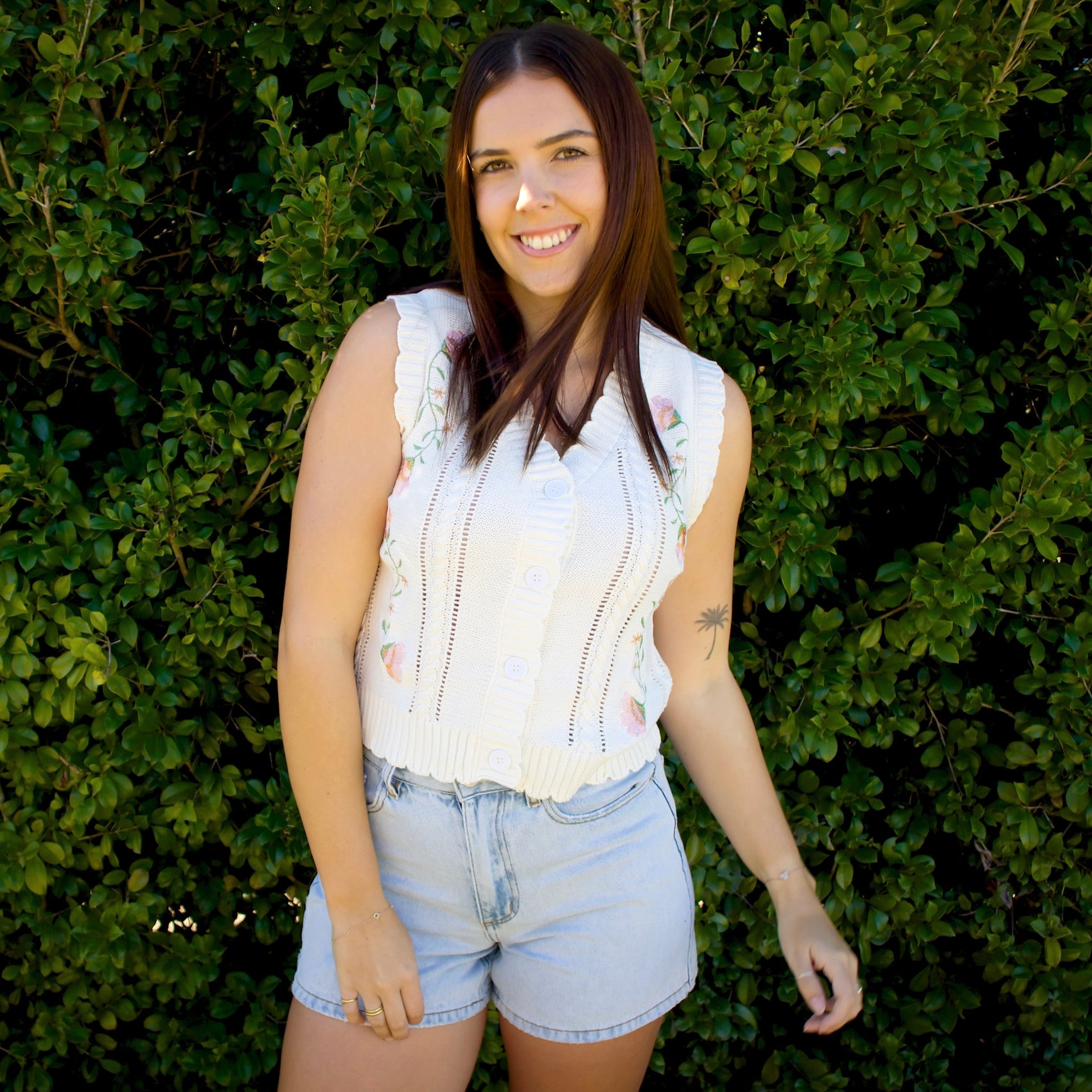 Woman wearing a white sleeveless top and denim shorts standing in front of green foliage.