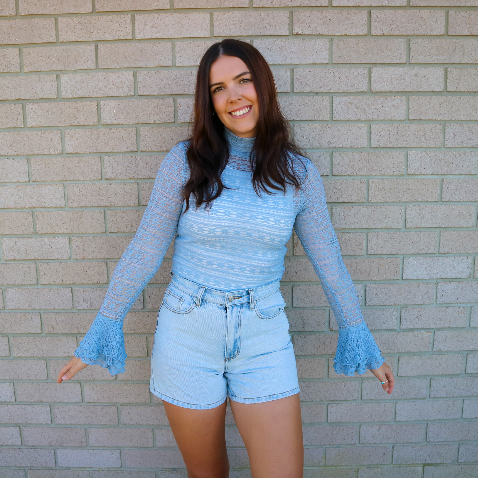 Woman wearing a blue lace top and denim shorts standing against a brick wall.