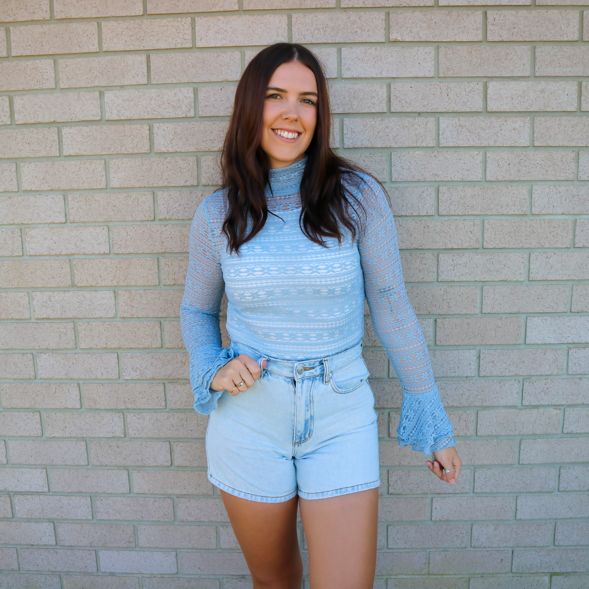 Woman wearing a light blue lace top and denim shorts standing against a brick wall.