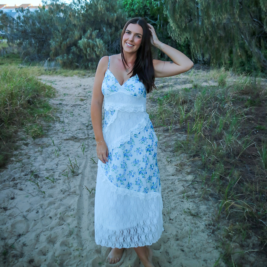 Woman in a white dress with blue floral pattern standing on a sandy path surrounded by greenery.