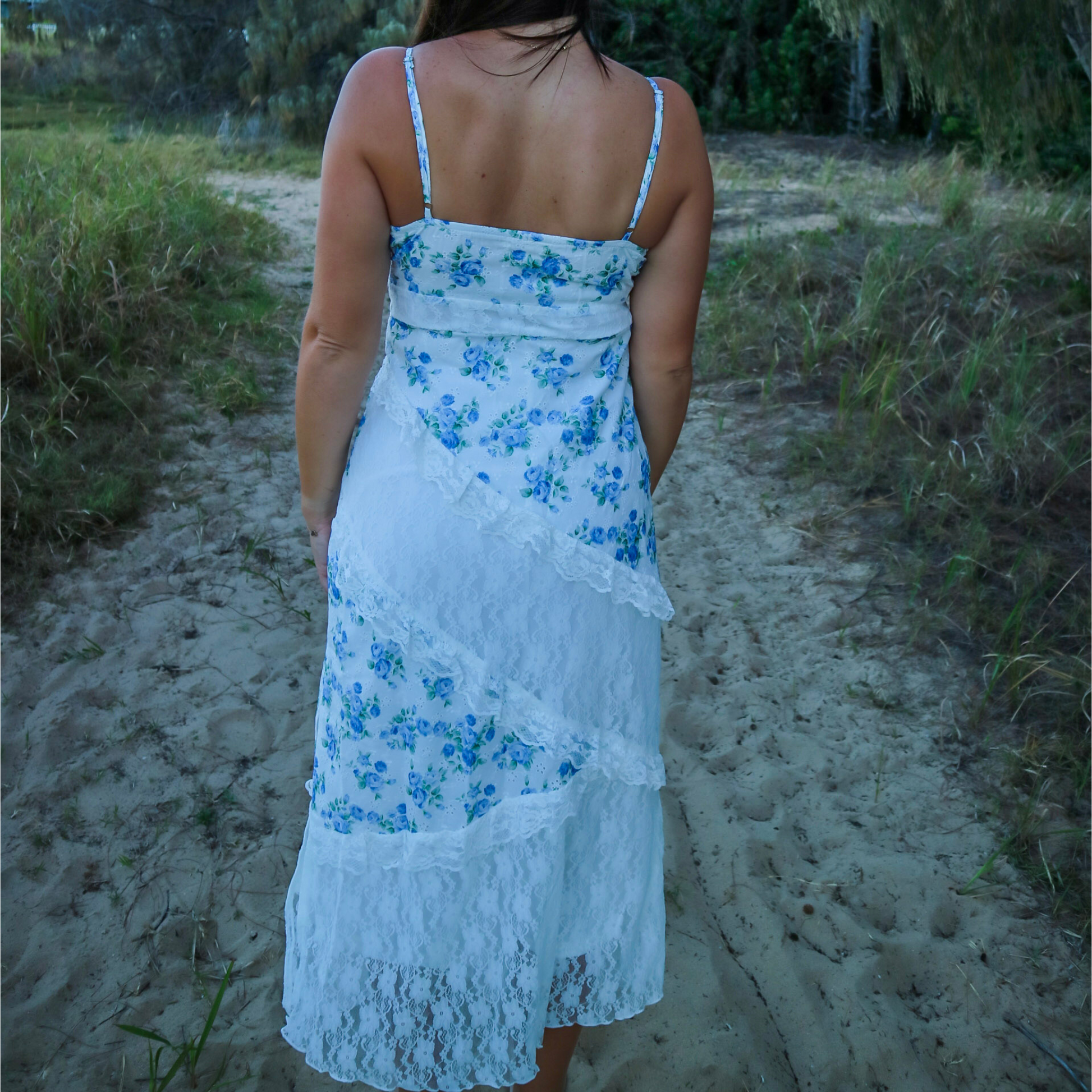 Woman wearing a white and blue floral dress standing on a sandy path with grass and trees in the background