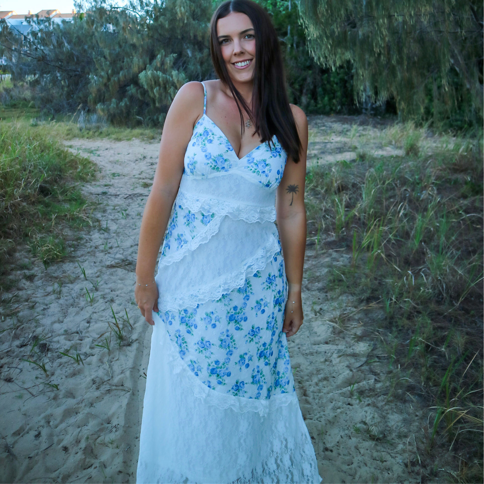 Woman in a white dress with blue floral patterns standing on a sandy path with greenery.