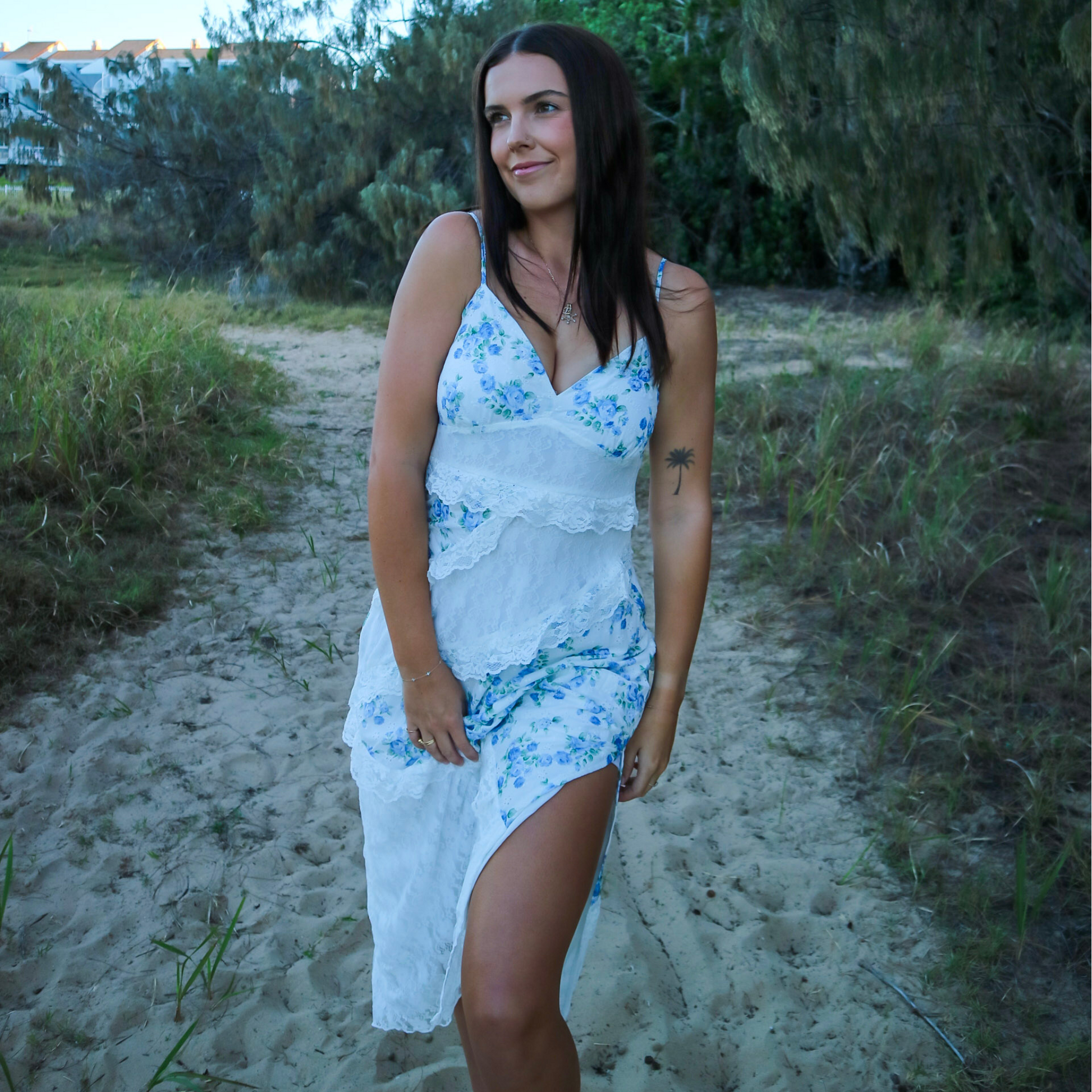 Woman in a white and blue floral dress standing on a sandy path with greenery around.