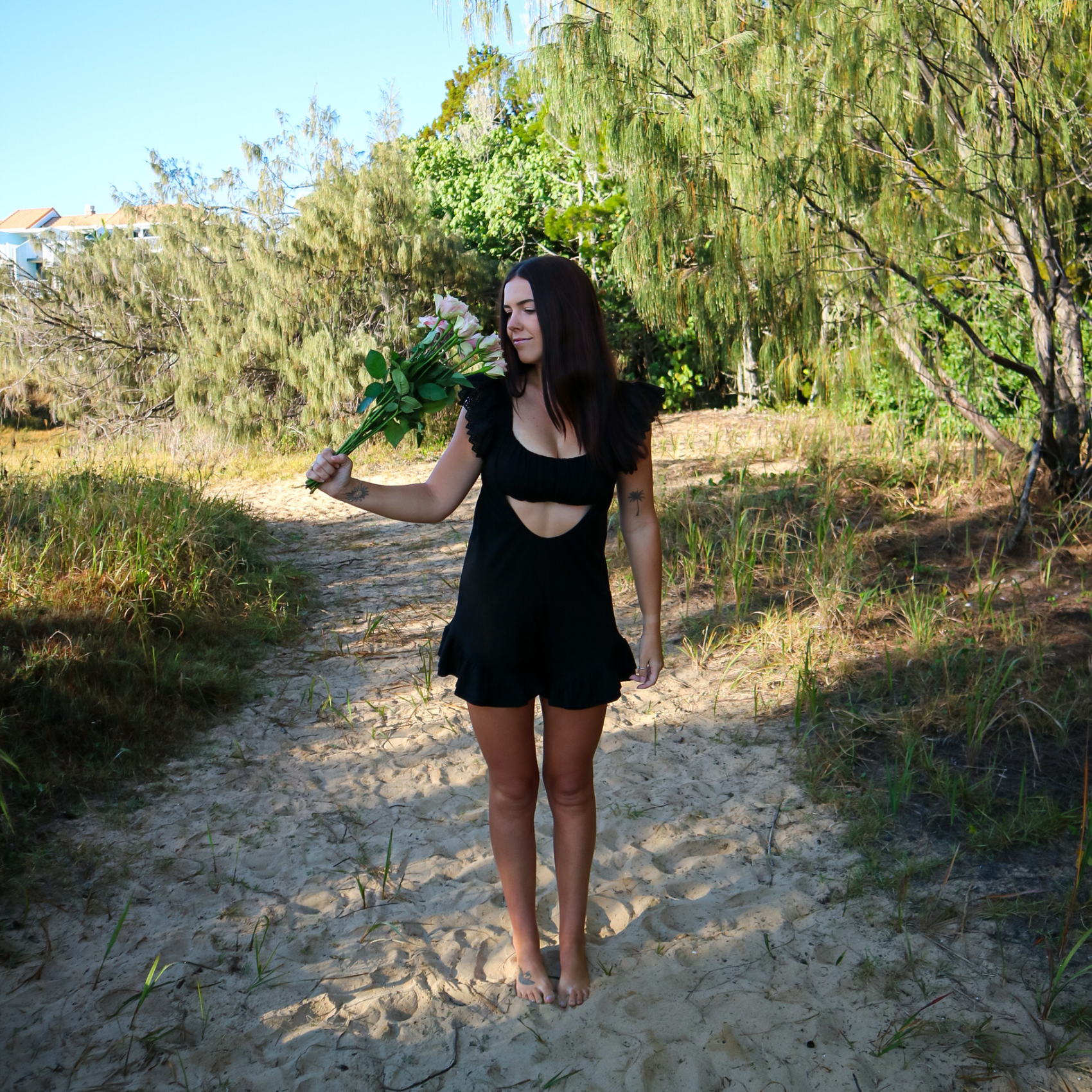 Woman in a black romper holding flowers on a sandy path with greenery