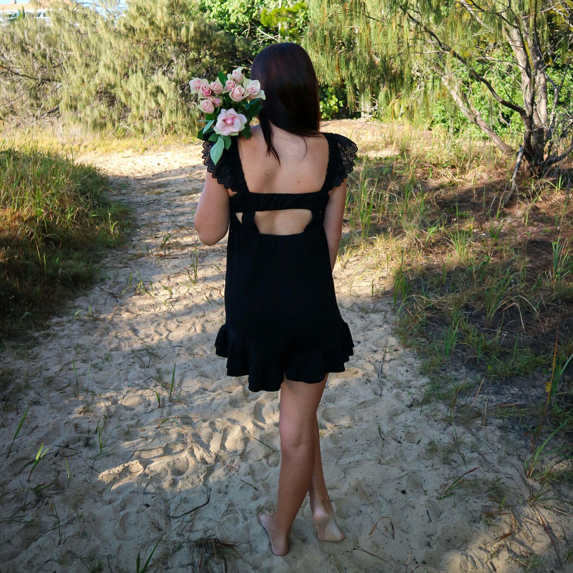 Woman in a black romper holding flowers on a sandy path with greenery