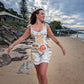 Woman in a patterned dress standing on a beach with rocks and trees in the background