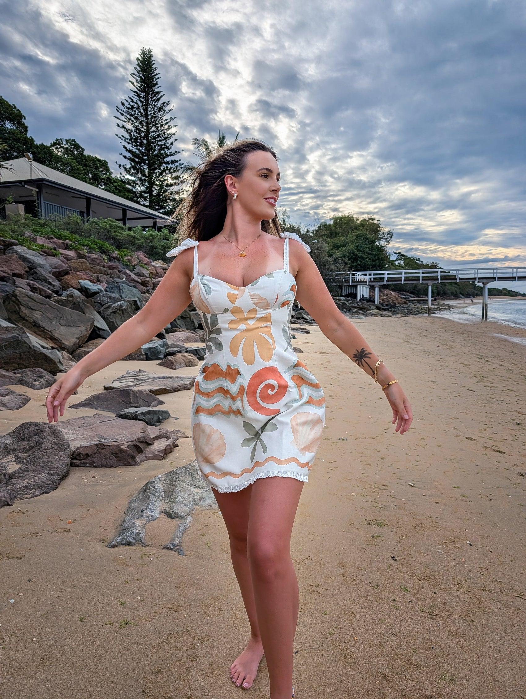 Woman in a patterned dress standing on a beach with rocks and trees in the background