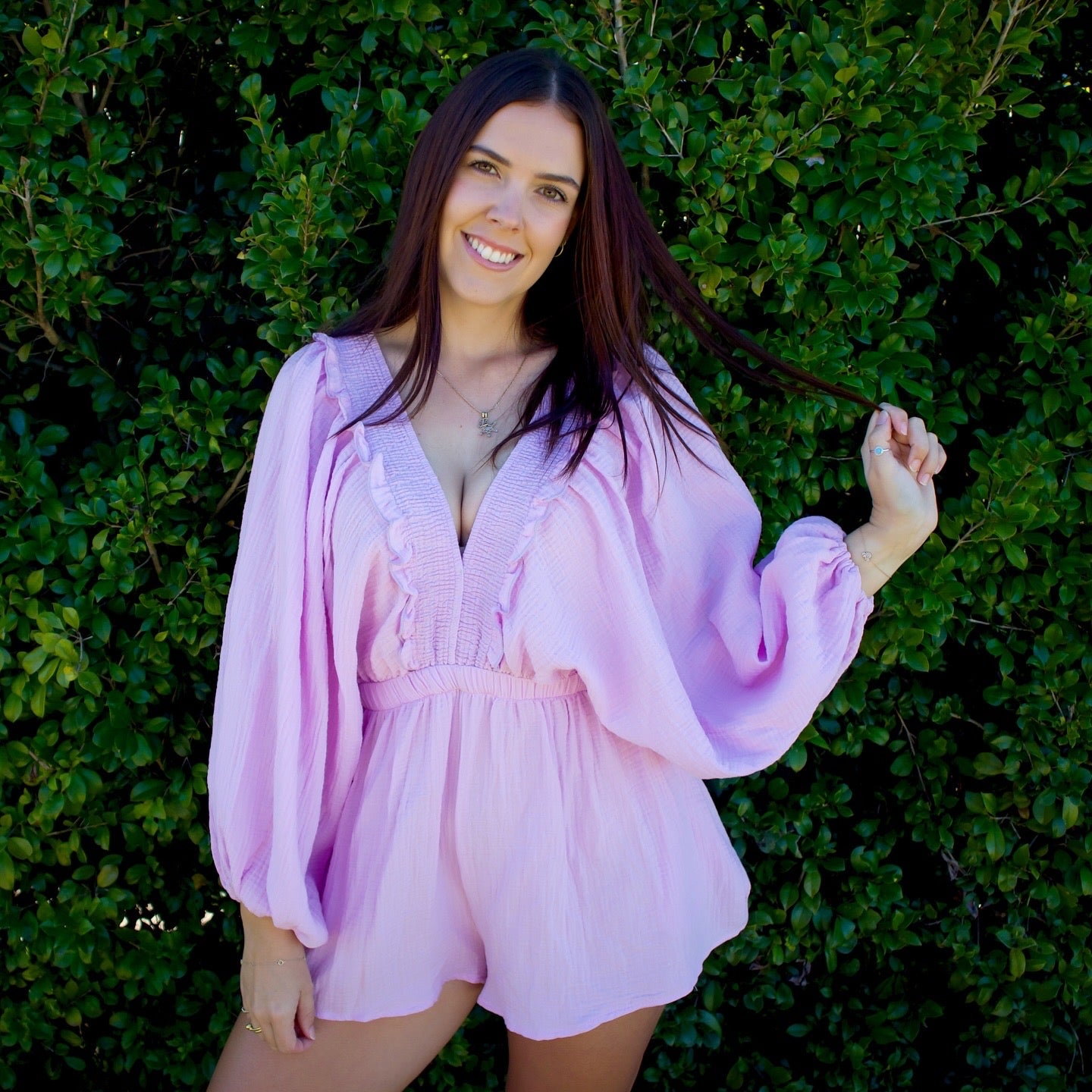 Woman wearing a purple romper standing in front of green foliage