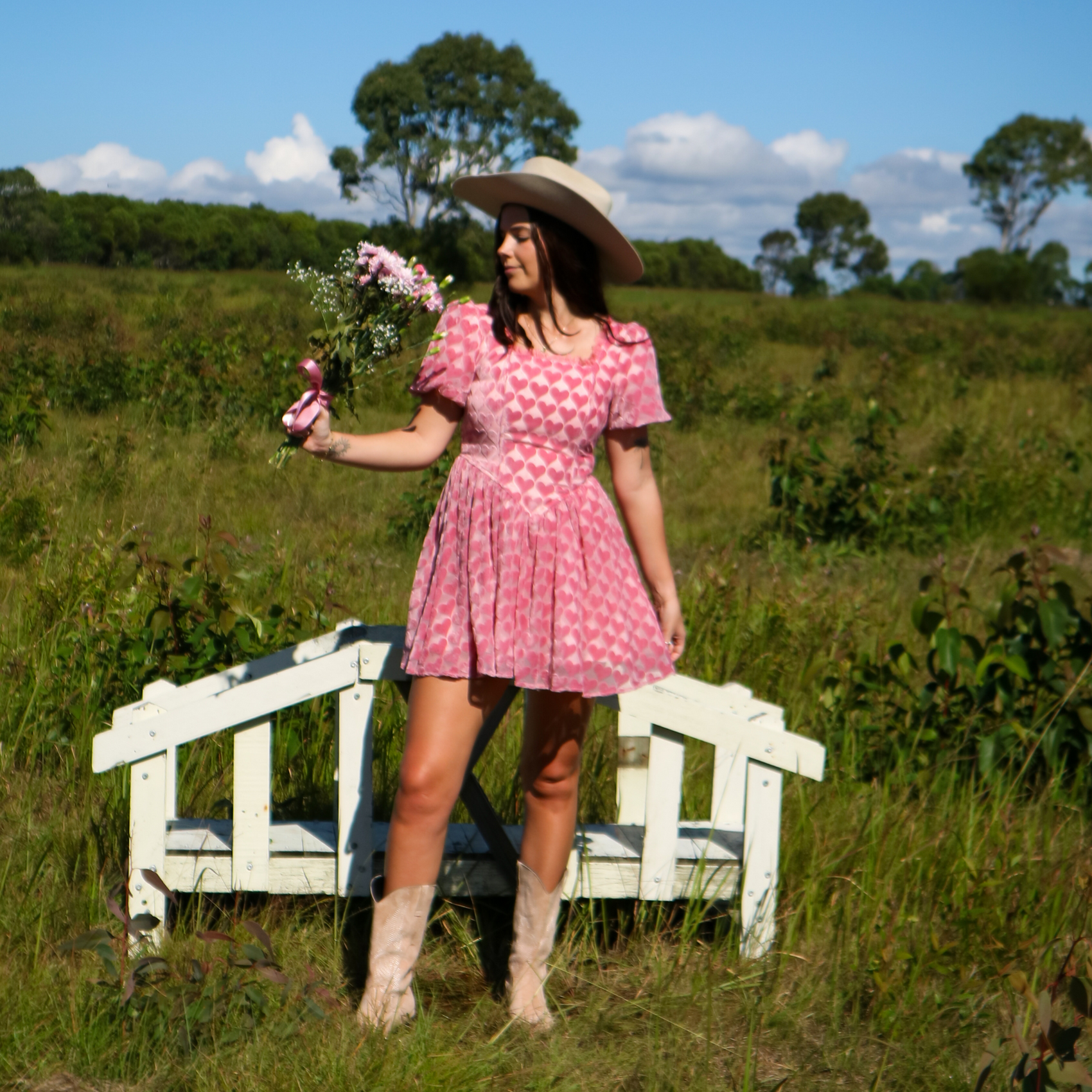 Woman in a pink dress and cowboy hat standing in a field with a white bench and flowers.