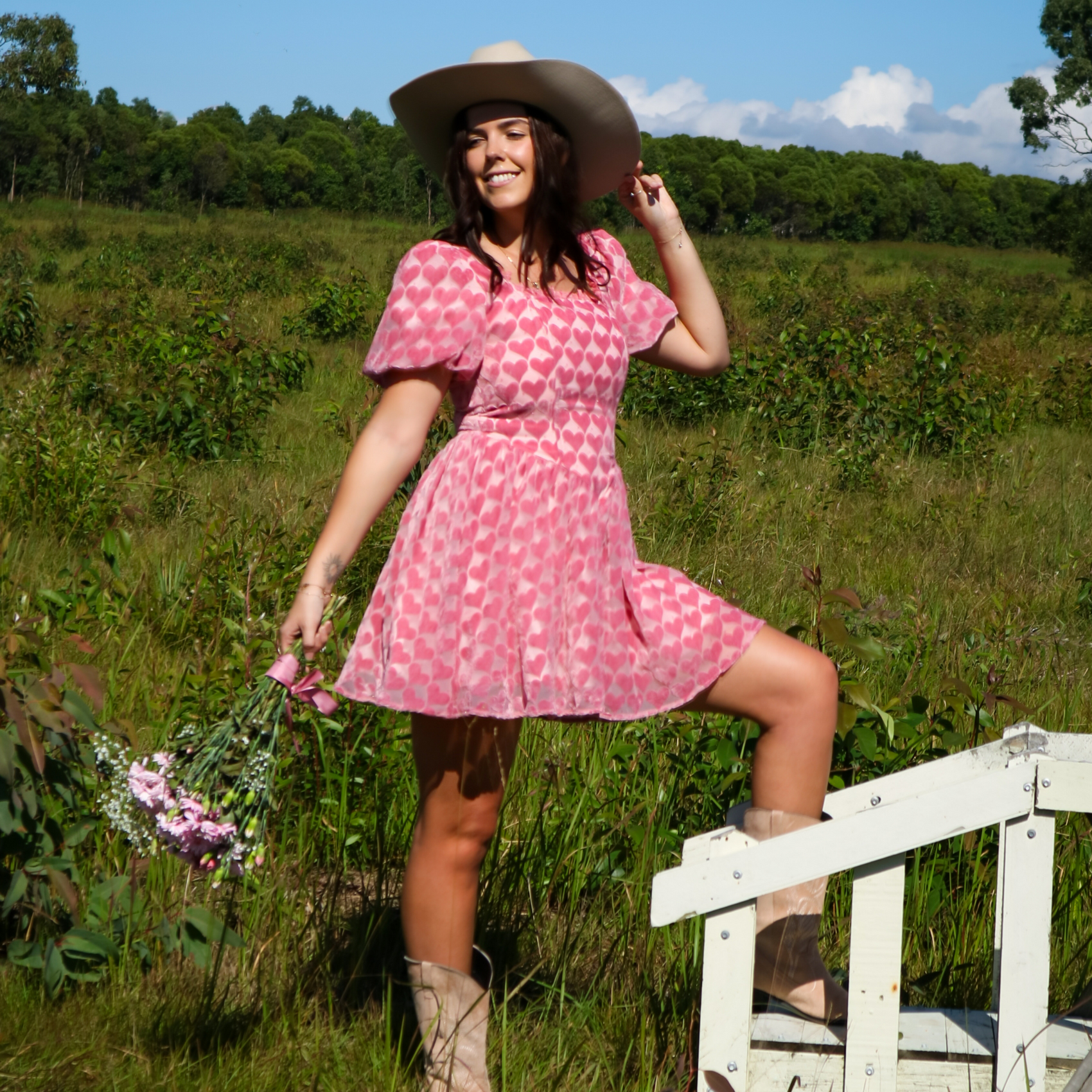 Woman in a pink heart dress and cowboy hat standing in a field with flowers.