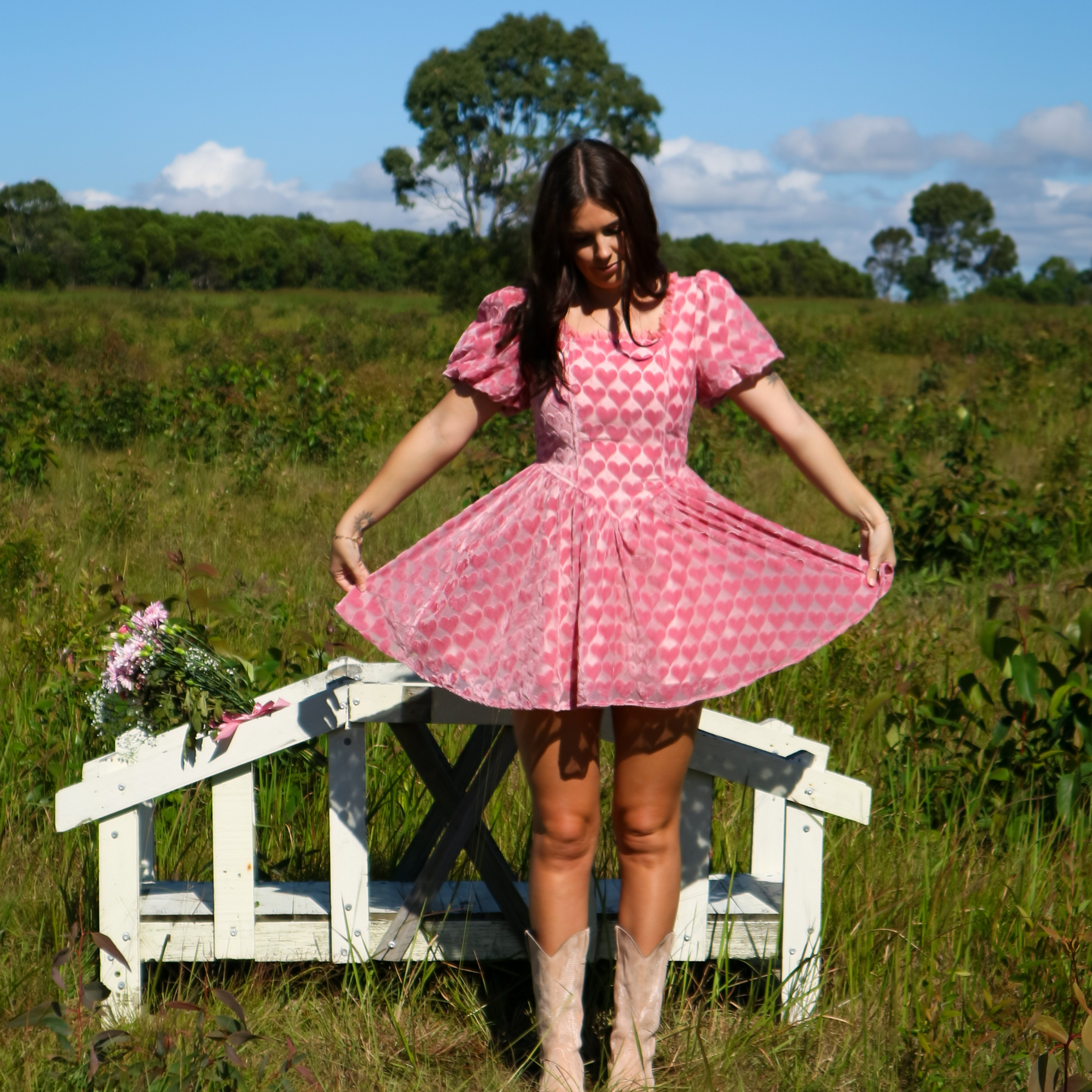 Woman in a pink heart dress standing in a field with a white bench and flowers.