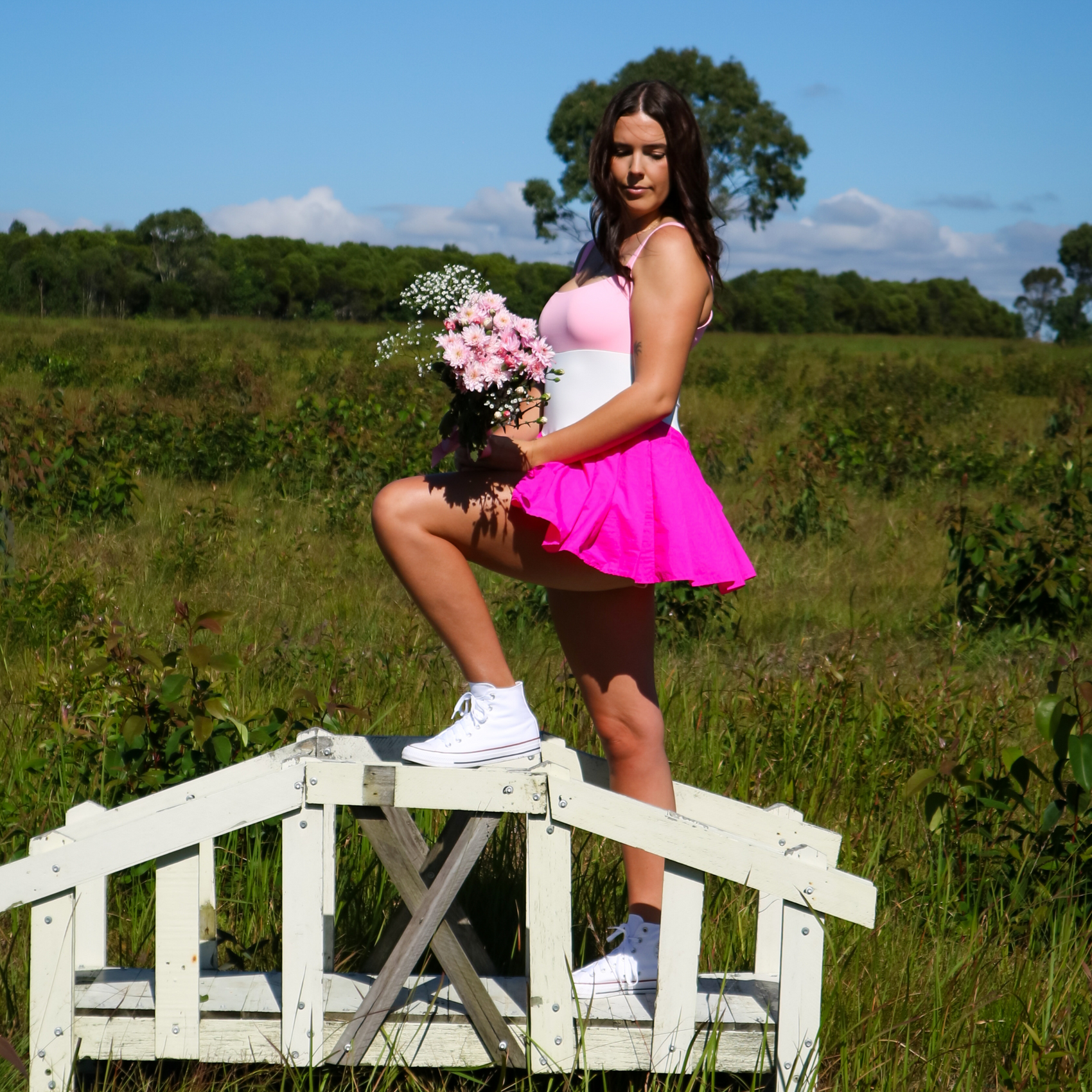 Woman in a pink dress standing on a wooden bridge in a field with flowers.