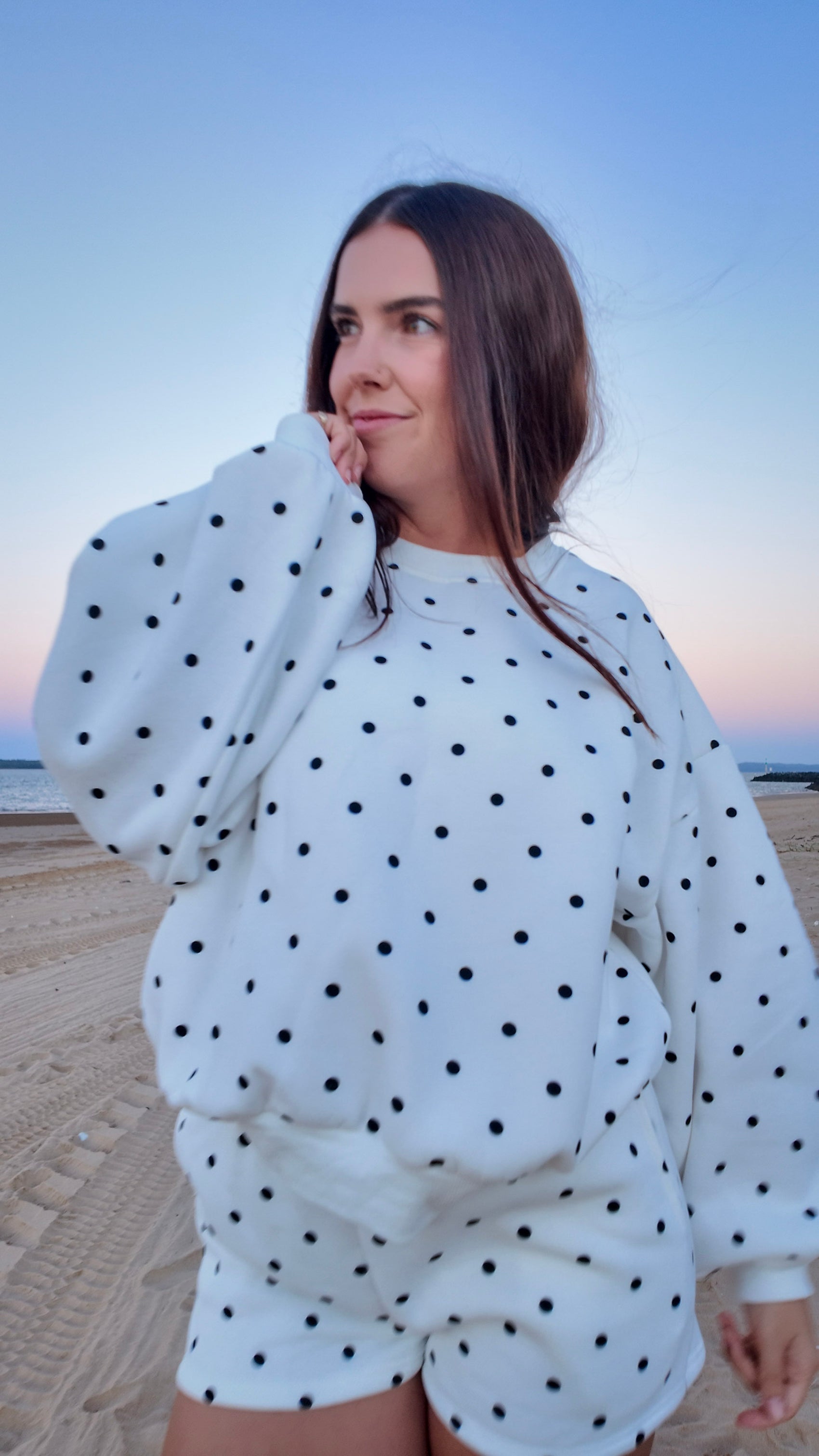 Woman wearing a white polka dot outfit standing on a beach at sunset.