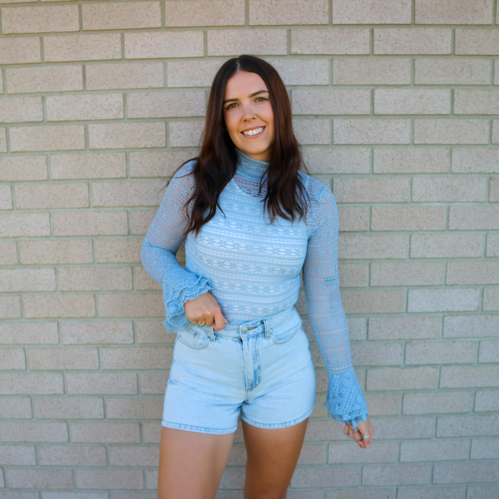 Woman wearing a blue turtleneck and denim shorts standing against a brick wall.