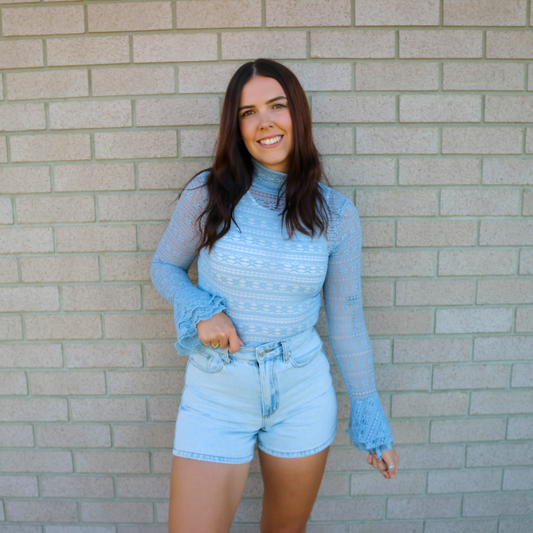 Woman wearing a blue turtleneck and denim shorts standing against a brick wall.