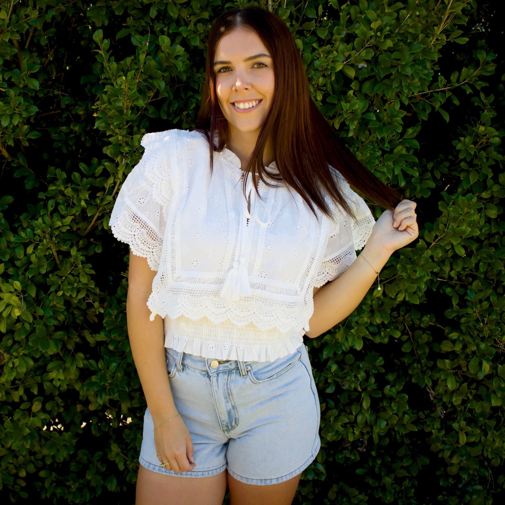 Woman wearing a white lace top and denim shorts standing against a green hedge.