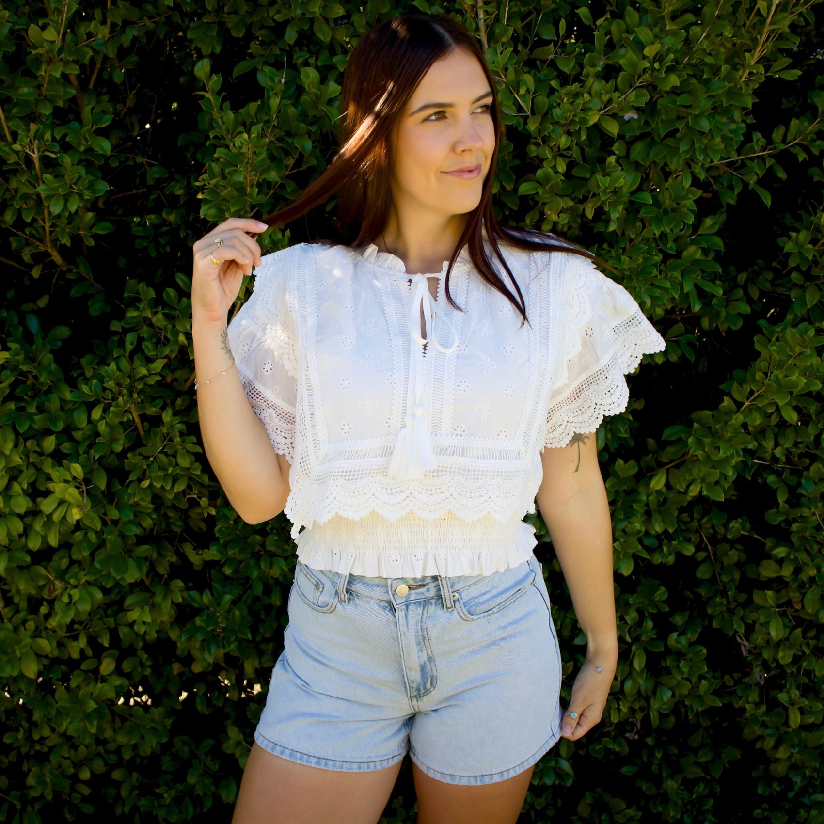 Woman wearing a white lace top and denim shorts standing against a green hedge.