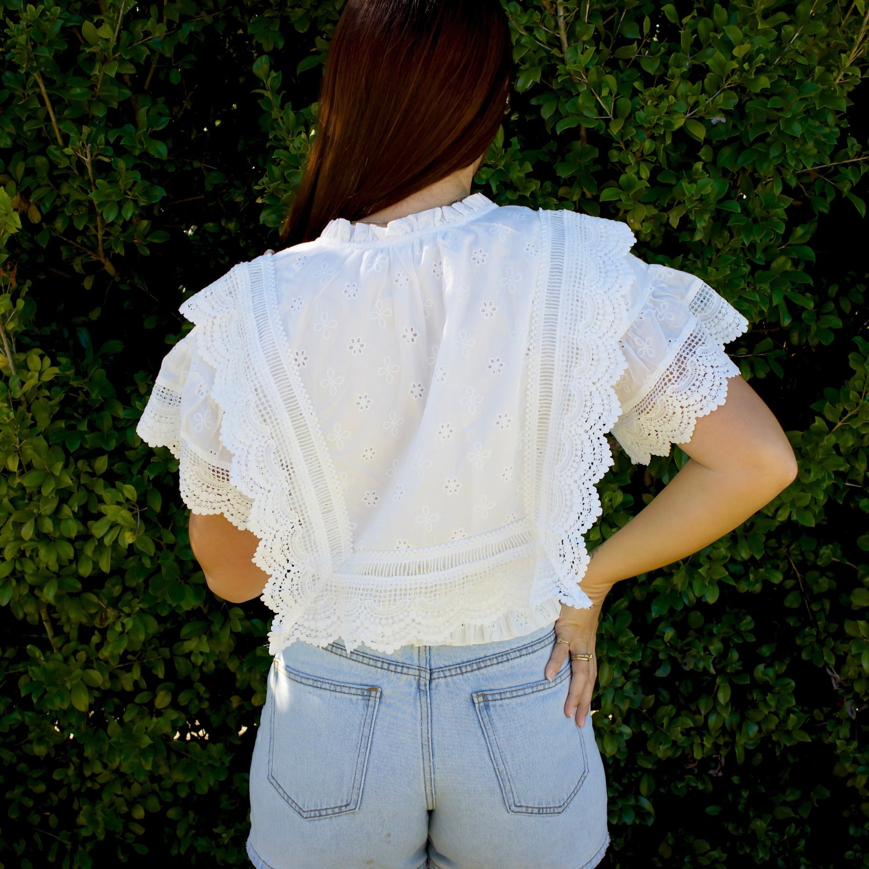 Person wearing a white lace top and light blue jeans against a green bush background