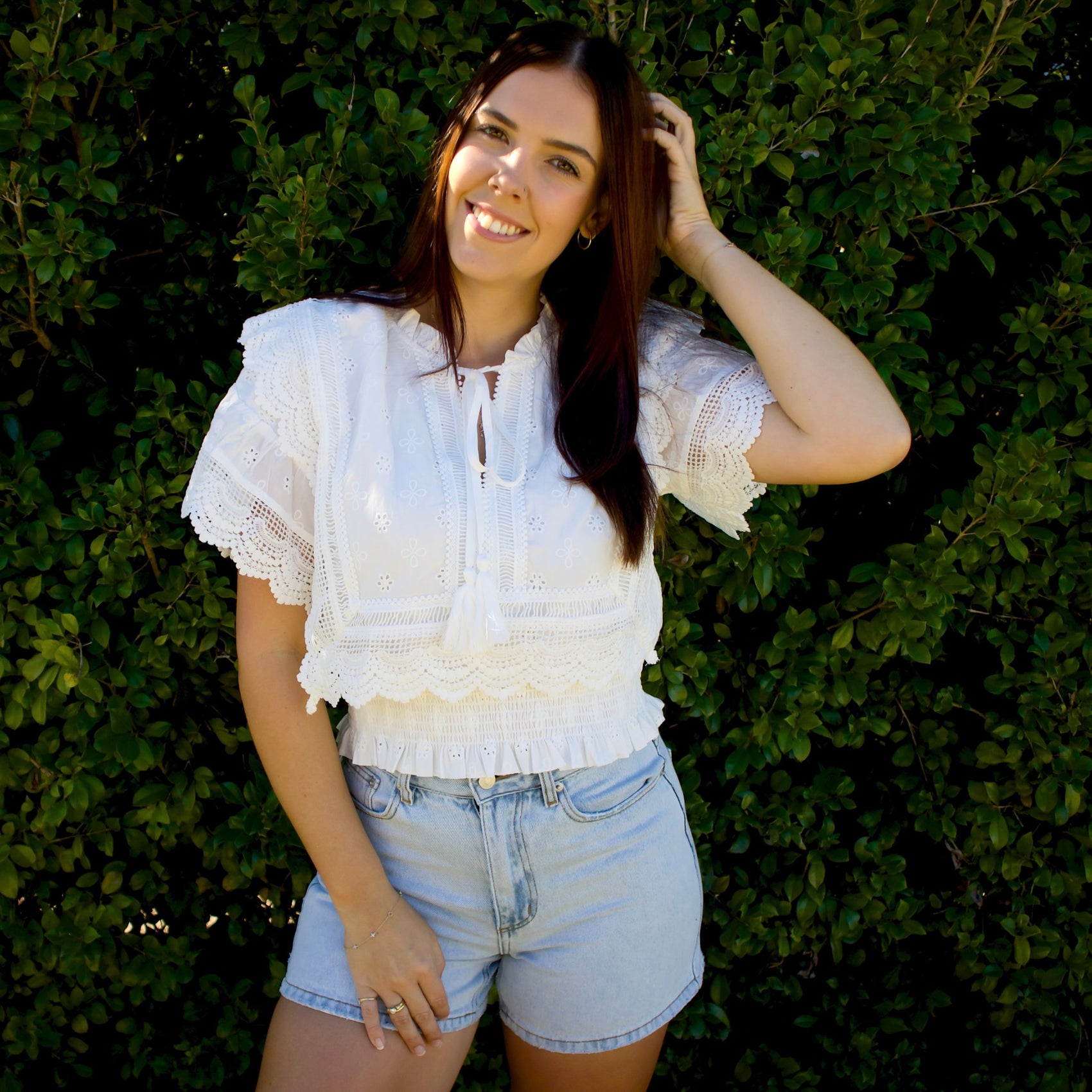 Woman wearing a white lace top and denim shorts standing against a green hedge.