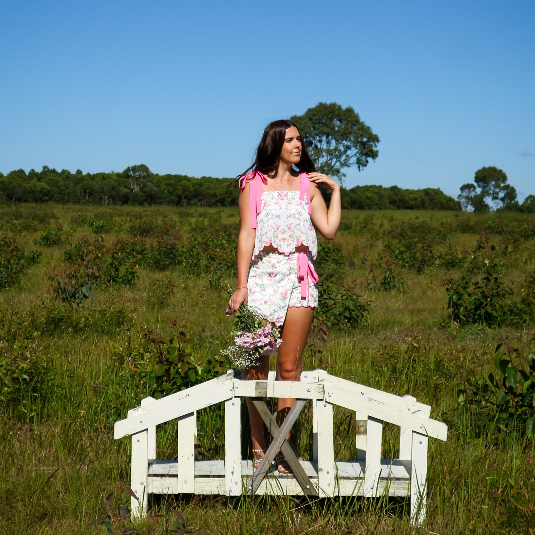 Woman standing on a small wooden bridge in a grassy field wearing a pink floral set.