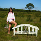 Woman standing on a small wooden bridge in a grassy field with a clear blue sky wearing a pink and white set.